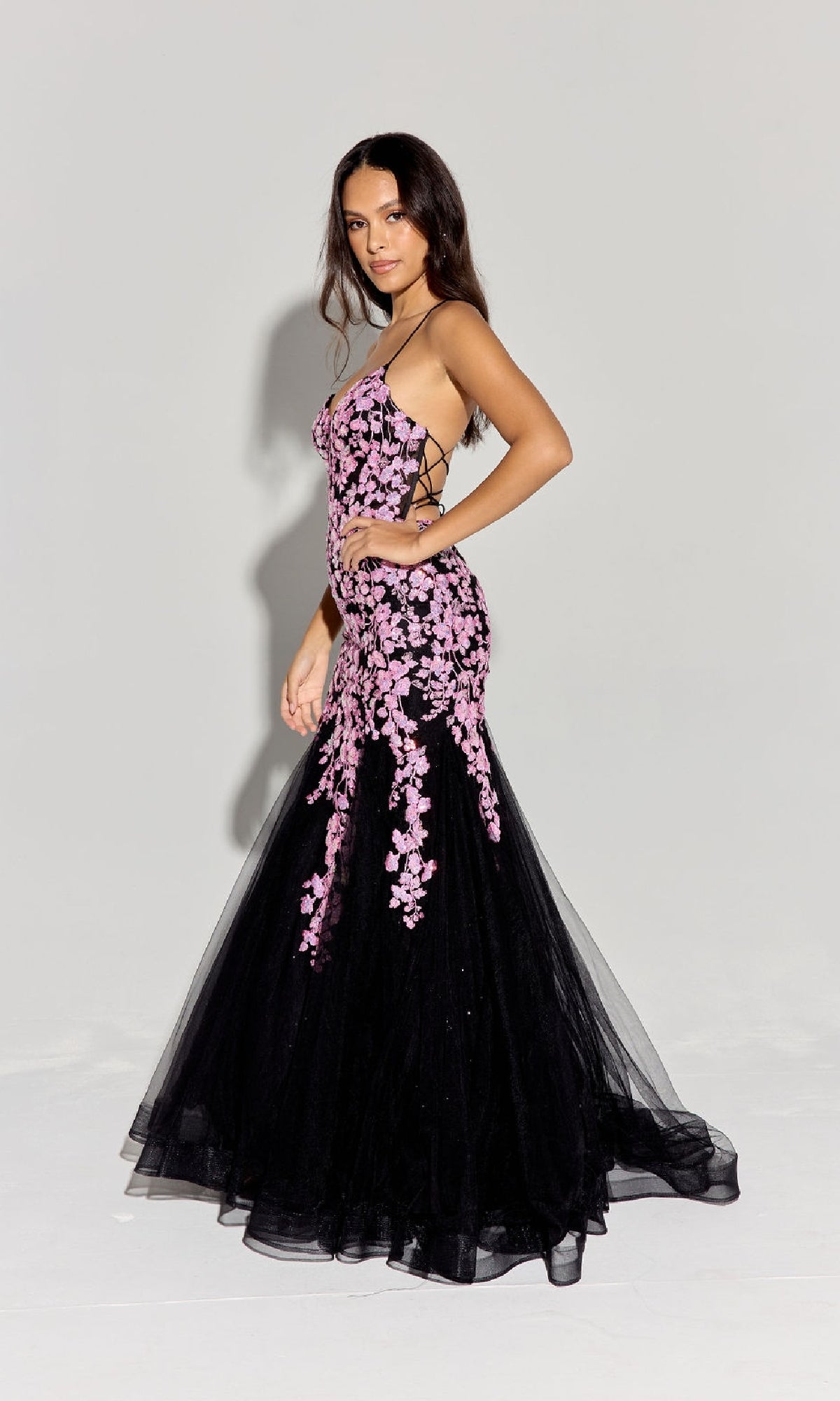 A woman poses in the Sequin-Floral Long Mermaid Prom Dress: Eureka 9696, featuring black fabric with pink floral embroidery and lace-up sides, against a plain light gray background.